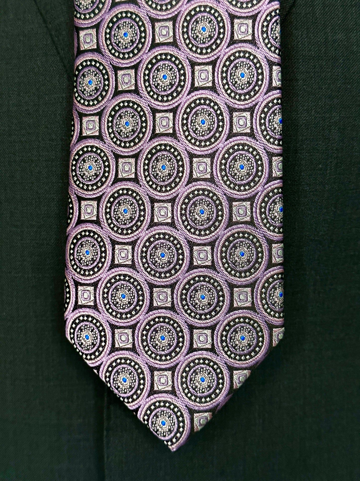 This 100% woven silk tie has a gorgeous weight and thickness to the finish. With woven medallions in shades of purple and black, this is a necktie that makes a statement. To be worn with shirts in white, blue, pink, beige and grey. This tie also pairs well with shirts with small plaids and mini checks. See the sister color in blue.