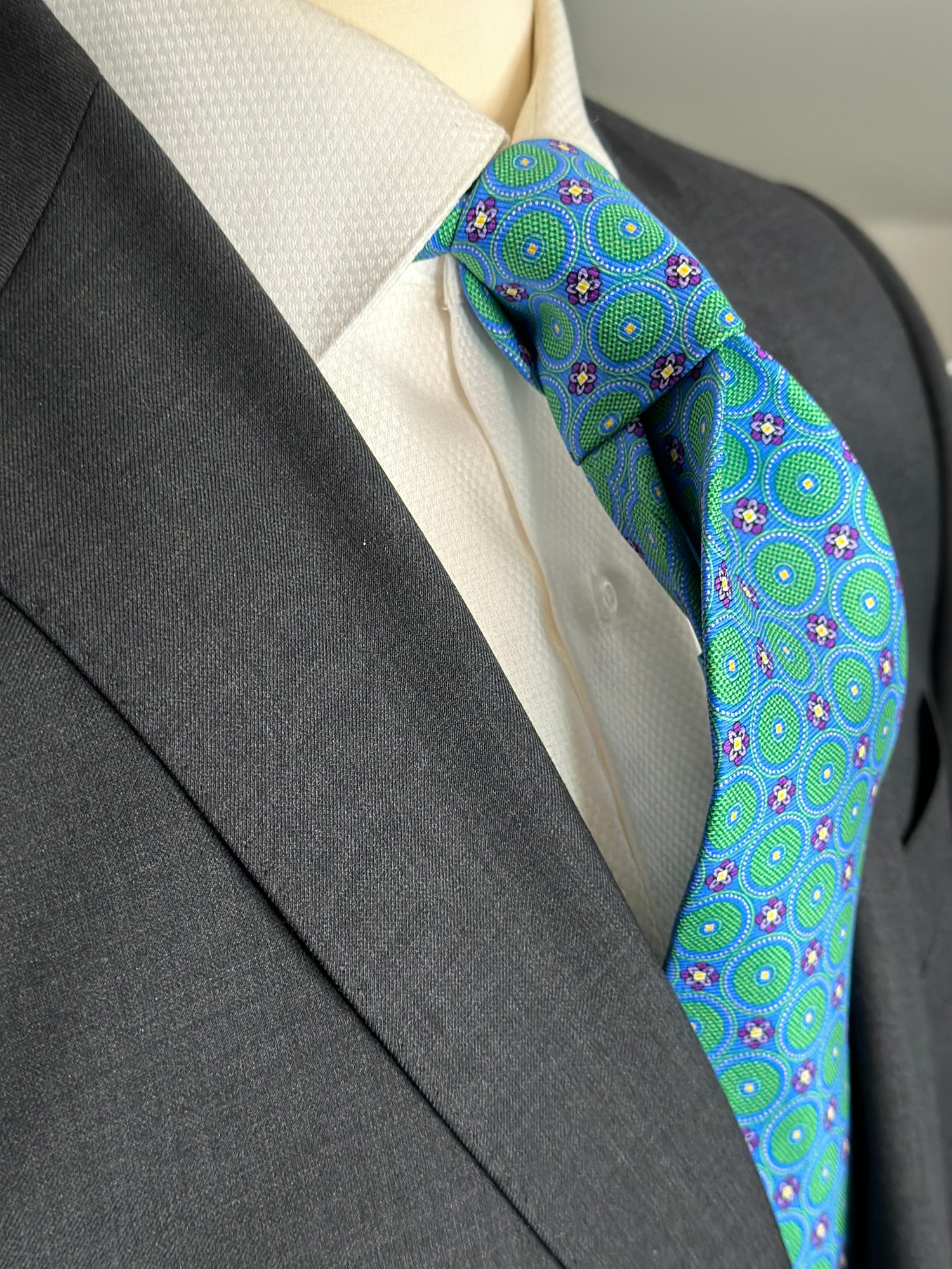 This tie with circular medallions in green surrounded by a beautiful blue background is 100% silk. Paired with dark suits and stripes this necktie allows for great soft contrast on white or sky blue and grey dress shirts. Try the sister tie in pink and blue for more combinations.