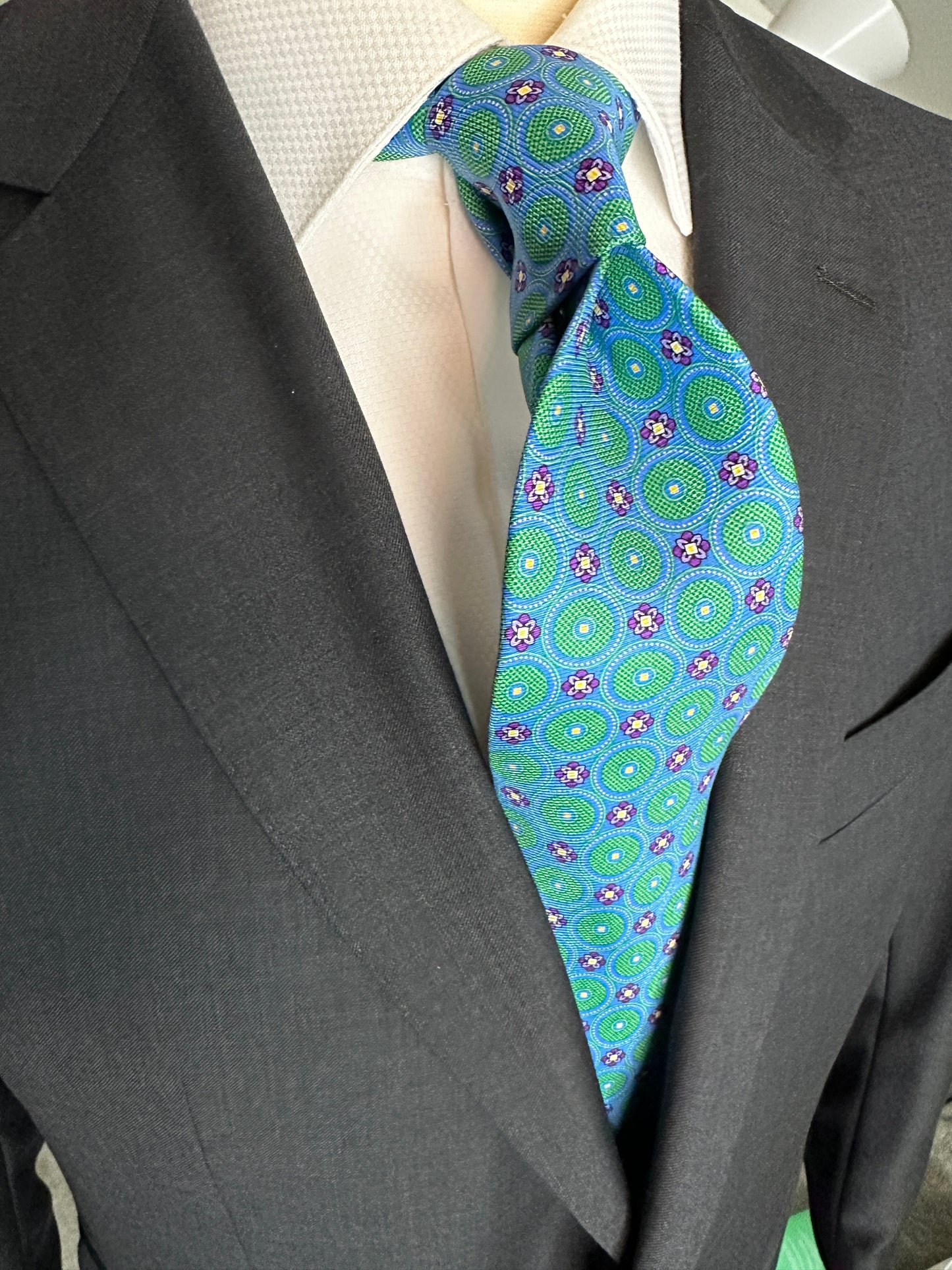 This tie with circular medallions in green surrounded by a beautiful blue background is 100% silk. Paired with dark suits and stripes this necktie allows for great soft contrast on white or sky blue and grey dress shirts. Try the sister tie in pink and blue for more combinations.