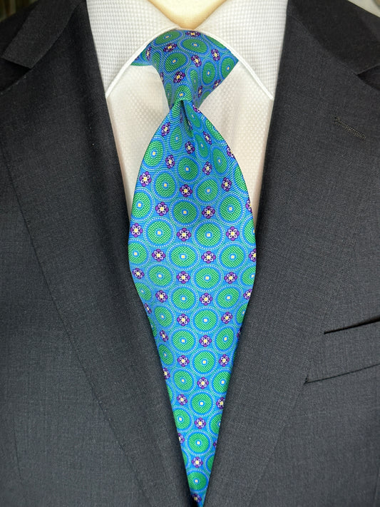 This tie with circular medallions in green surrounded by a beautiful blue background is 100% silk. Paired with dark suits and stripes this necktie allows for great soft contrast on white or sky blue and grey dress shirts. Try the sister tie in pink and blue for more combinations.