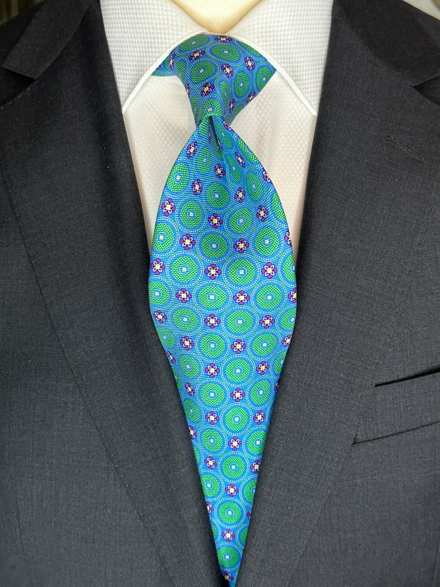 This tie with circular medallions in green surrounded by a beautiful blue background is 100% silk. Paired with dark suits and stripes this necktie allows for great soft contrast on white or sky blue and grey dress shirts. Try the sister tie in pink and blue for more combinations.