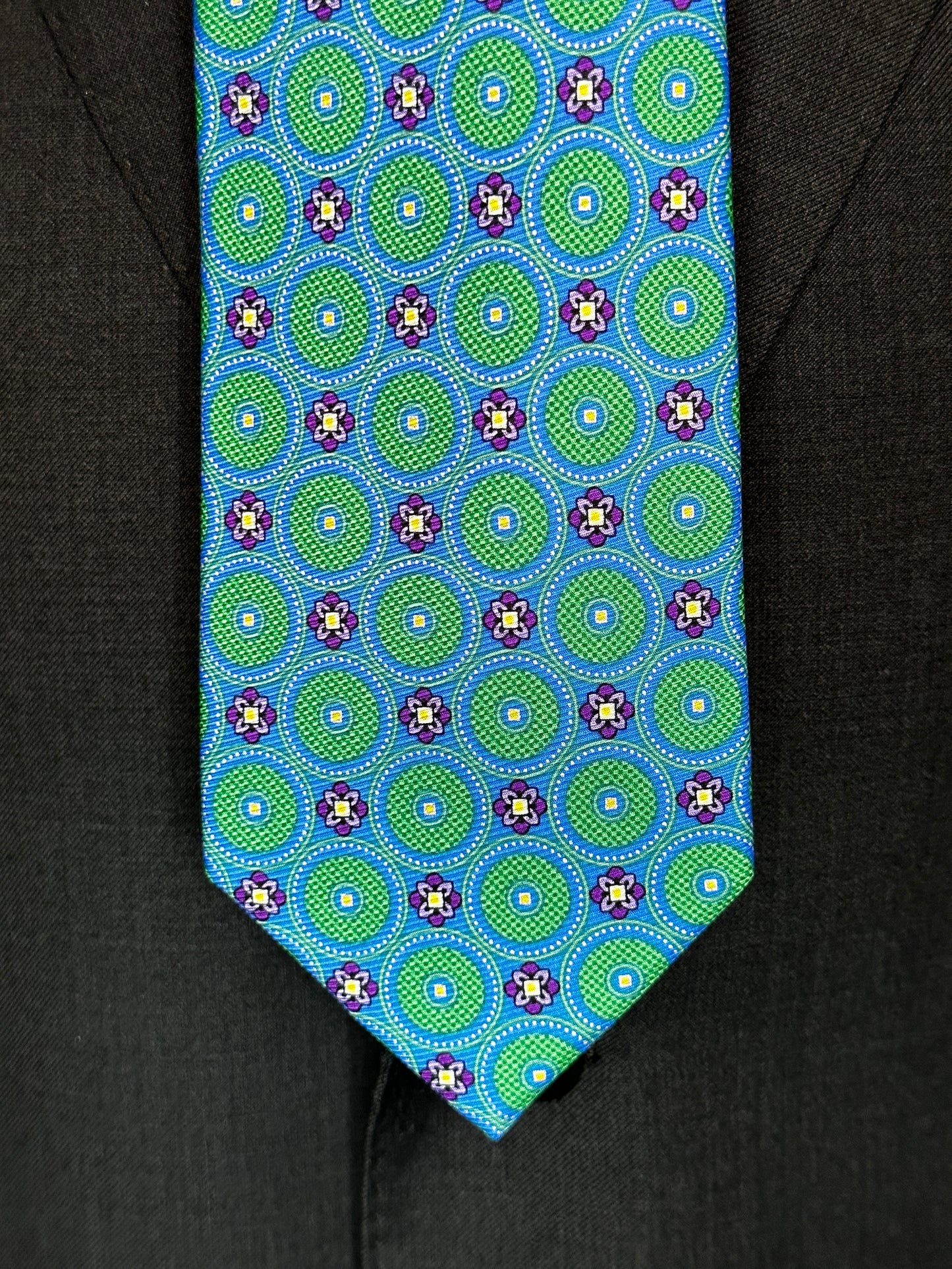 This tie with circular medallions in green surrounded by a beautiful blue background is 100% silk. Paired with dark suits and stripes this necktie allows for great soft contrast on white or sky blue and grey dress shirts. Try the sister tie in pink and blue for more combinations.