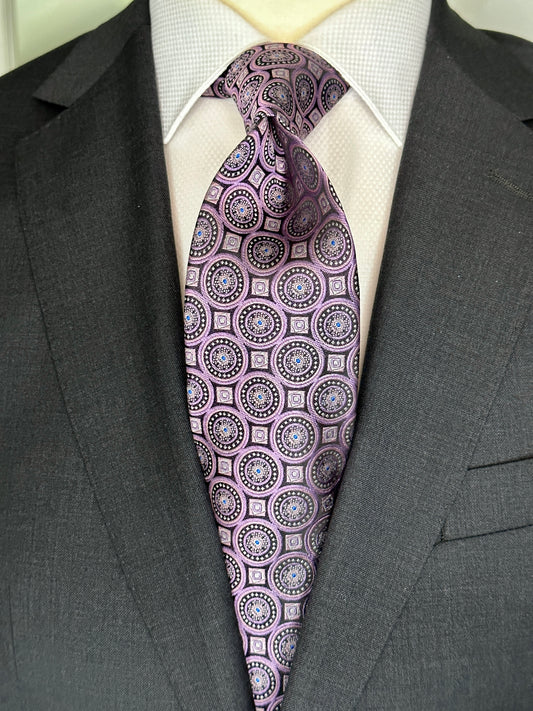 This 100% woven silk tie has a gorgeous weight and thickness to the finish. With woven medallions in shades of purple and black, this is a necktie that makes a statement. To be worn with shirts in white, blue, pink, beige and grey. This tie also pairs well with shirts with small plaids and mini checks. See the sister color in blue.