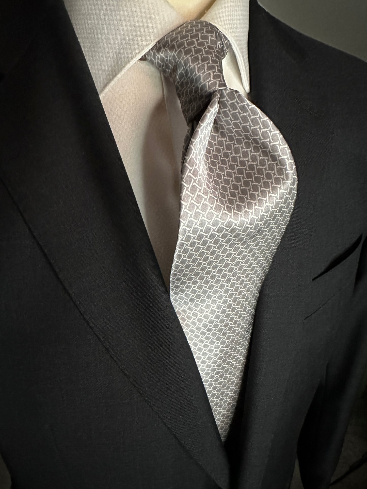 This satin finish 100% silk necktie has a very interesting all over geometric pattern of interlocking white links almost chain link in style. The silk is very smooth to the touch and has a natural luster that shines in certain light. This is a serious tie. Great for those business meetings, court dates, daytime or evening events, and all around more formal attire. This is best paired with a fancy white or tone on tone white dress shirt.